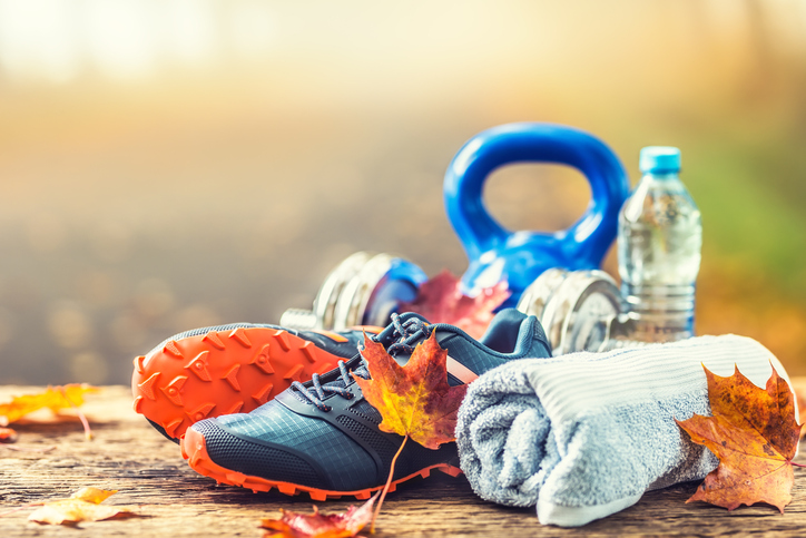 Pair of blue sport shoes, water and dumbbells laid on a wooden board in a tree autumn alley with maple leaves - accessories for run exercise or workout activity.